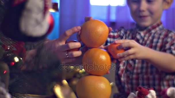 Family Party Happy Kid Playing with Oranges Mother Plays With Son Image is Blurred Then Focused on Red and Gilded Christmas Balls and Bows Evening Indoors — стоковое видео