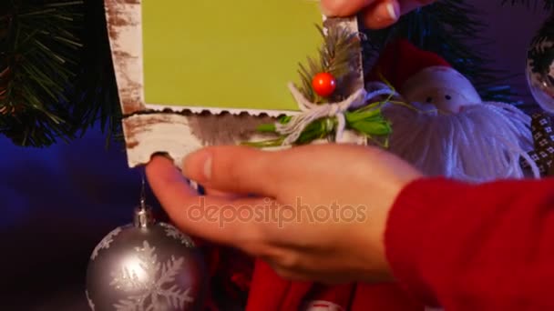 Des mains masculines prennent une poche faite à la main avec un cadre photo sous un sapin de Noël décoré, avec beaucoup de boules scintillantes allongées sur un sol violet — Video
