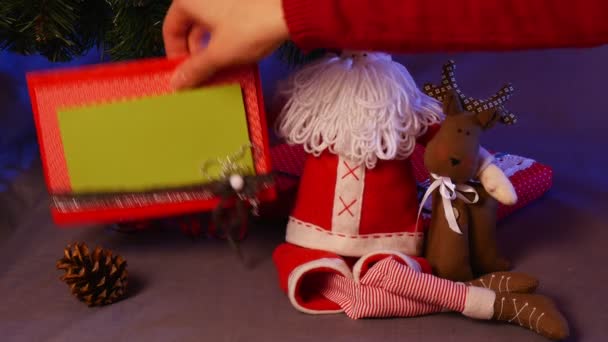 Moldura de foto feita à mão é tirada e colocada de volta sob uma árvore de Natal com um monte de brinquedos engraçados sob ele, incluindo um veado marrom, Papai Noel vermelho, cones e bolas — Vídeo de Stock