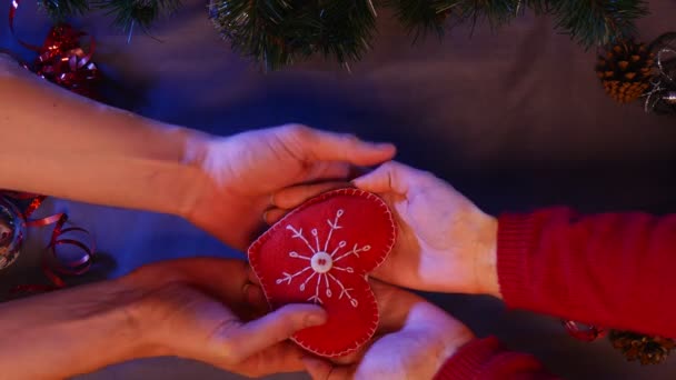 Un bonito corazón rojo hecho a mano se entrega debajo del árbol de Navidad con conos de pino y rayas de lluvia de colores hechos a mano en las inmediaciones — Vídeos de Stock