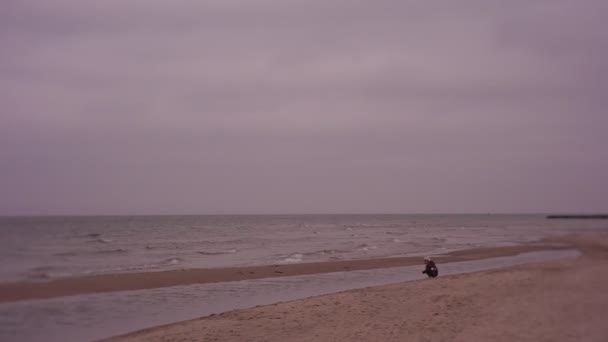 Κοριτσάκι κάθεται στην παραλία και είναι σε αναμονή, ένα παιδί το χειμώνα στη θάλασσα — Αρχείο Βίντεο