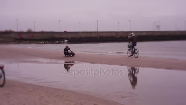 Par på cyklar Rider förbi mamma och flickor leker med Sand på stranden, vintern havet spel — Stockvideo