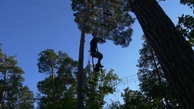 Tourist Silhouette on Rope Road, Man in Park of Amusement, Slow Motion. Safety Belts Are Fixed With Carabiners to the Cables. Male Climber in Adventure Rope Park in Summer Sunny Day