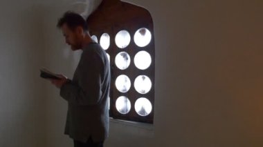 Elinde İncil olan bir adam pencerenin önünde. Believer yüksek sesle okuyor, odanın yanından geçiyor ve meditasyon yapıyor. Ortodoks Kilisesi, Manastır. Küçük tahta pencere çerçevesi. Ahşap Kutulu Mağara Açısı