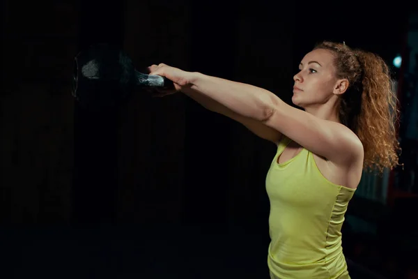 Mulher fitness balançando o sino da chaleira no ginásio. Mulher caucasiana jovem fazendo exercício de balanço com um kettlebell como uma rotina de um treino crossfit . — Fotografia de Stock