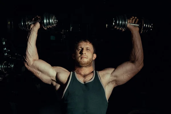 Homem bonito com bíceps grandes fazendo exercícios com halteres em um fundo preto. Fisiculturista — Fotografia de Stock