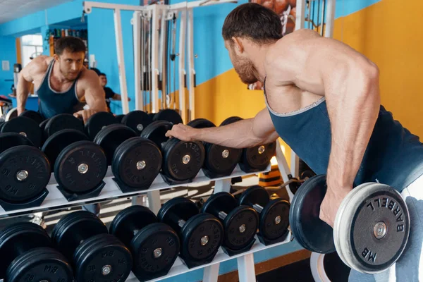 Um grande e bonito fisiculturista forte do sexo masculino fazendo um supino no espelho, faz um grande bíceps usando um haltere no ginásio . — Fotografia de Stock