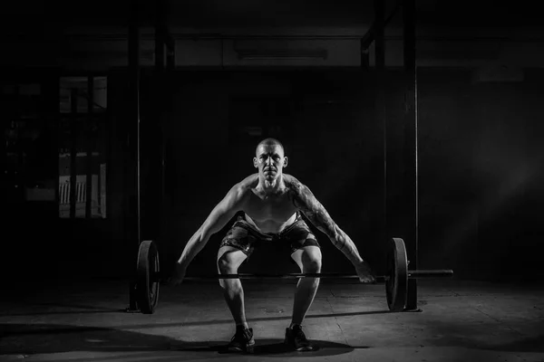 En muskulös man knäböj med en skivstång på gymmet. Svartvitt foto — Stockfoto
