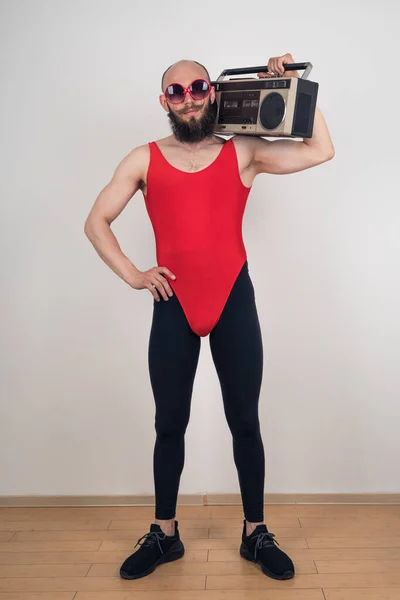 A man dressed for disco-retro parties. Funny bearded guy in sunglasses and leggings with red bodysuit with retro tape recorder