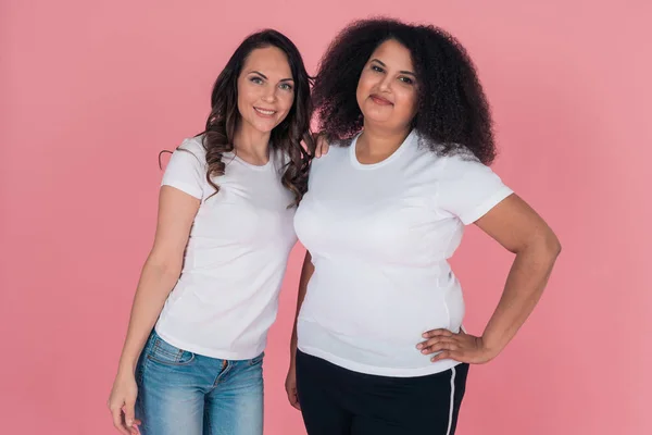 Twee leuke meisjes een Afro-Amerikaanse en een blank meisje glimlachen direct naar de camera in witte t-shirts op een roze achtergrond — Stockfoto