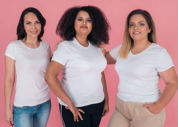 Drie vriendinnen lachen prachtig en poseren voor de camera op een roze achtergrond. Meisjes in witte t-shirts. Uitvoeringen voor ontwerp — Stockfoto
