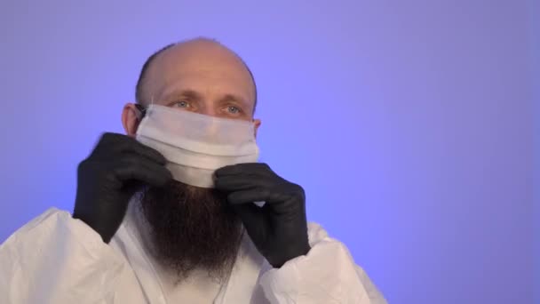 Hombre caucásico barbudo en un traje protector blanco y guantes negros con una máscara, gafas y capucha . — Vídeos de Stock