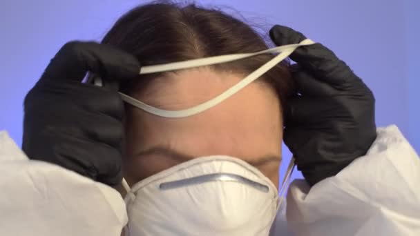 Donna caucasica in tuta protettiva bianca su uno sfondo blu mette sul suo viso una maschera medica, occhiali e indossa un cappuccio . — Video Stock