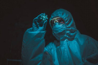 Coronavirus kırmızı modelini elinde tutan koruyucu kıyafetli bir kadın bilim adamı. COVID-19 molekülü olan beyaz bir kadın..