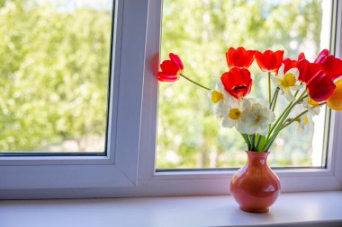 Beyaz bir pencere pervazında vazoda duran parlak kırmızı, beyaz ve sarı çiçekler. Pencerenin dışında ağaçların yeşil yaprakları var. Pencereden güneş ışığı düşüyor.