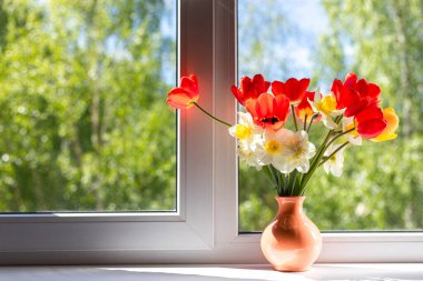 Beyaz bir pencere pervazında vazoda duran parlak kırmızı, beyaz ve sarı çiçekler. Pencerenin dışında ağaçların yeşil yaprakları var. Pencereden güneş ışığı düşüyor.