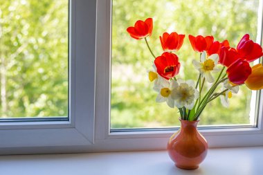 Beyaz bir pencere pervazında vazoda duran parlak kırmızı, beyaz ve sarı çiçekler. Pencerenin dışında ağaçların yeşil yaprakları var. Pencereden güneş ışığı düşüyor.