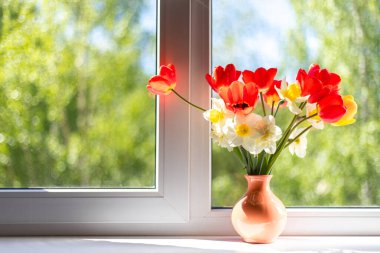 Beyaz bir pencere pervazında vazoda duran parlak kırmızı, beyaz ve sarı çiçekler. Pencerenin dışında ağaçların yeşil yaprakları var. Pencereden güneş ışığı düşüyor.