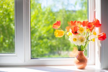 Beyaz bir pencere pervazında vazoda duran parlak kırmızı, beyaz ve sarı çiçekler. Pencerenin dışında ağaçların yeşil yaprakları var. Pencereden güneş ışığı düşüyor.