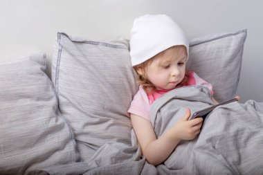 Beyaz şapkalı bebek yatakta akıllı telefon çalıyor.