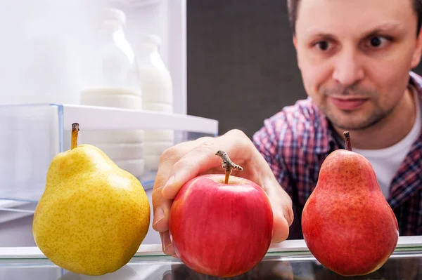 Man in a checked shirt takes red apple out from refrigerator inside look