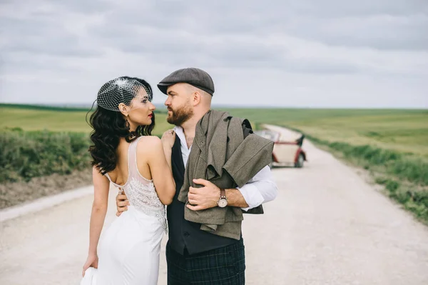 Elegante pareja de boda, novia, novio caminando en un campo nea carretera —  Fotos de Stock