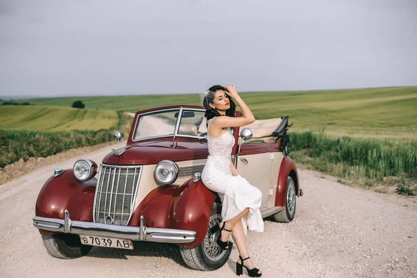 Noiva bonita elegante sentado em um carro retro vermelho em uma estrada de campo . — Fotografia de Stock
