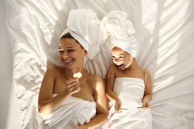 Beyaz banyo havlularına sarılı beyaz saçlarıyla genç bir beyaz anne ve küçük kızı yatağa uzanmış ve gözlerine salatalık parçaları sürüyorlar. Aile kaplıcası. Üst görünüm