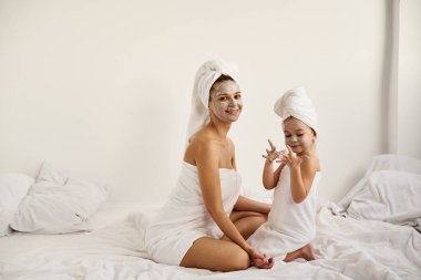 Beyaz banyo havlularına sarılı beyaz saçlı beyaz bir anne ve küçük kız anne yüzüne kilden bir maske takıyor.