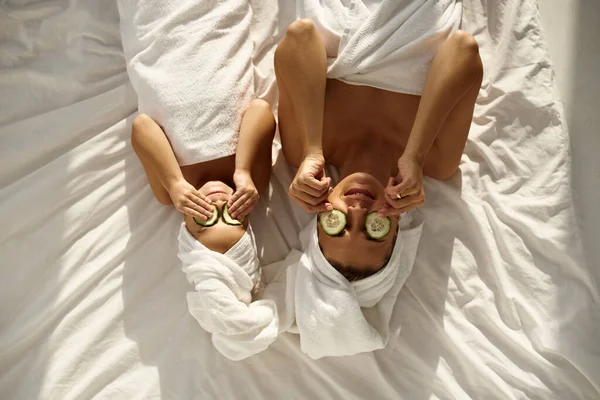 A young caucasian mother and little daughter with wrapped hair in white bath towels lying on the bed, and applying pieces of cucumber to their eyes. Family spa. Top view