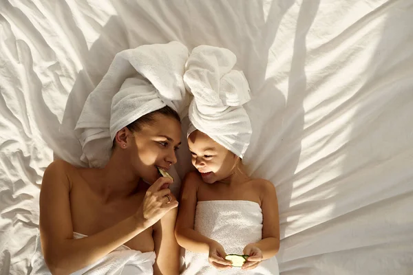 A young caucasian mother and little daughter with wrapped hair in white bath towels lying on the bed, and applying pieces of cucumber to their eyes. Family spa. Top view