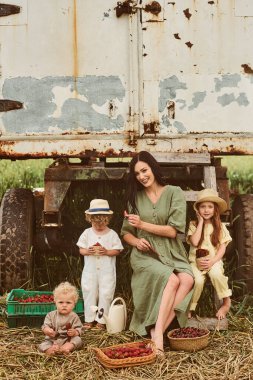 Bir sepet çilekle keten elbiseli güzel beyaz bir anne yeni bir mahsul topluyor ve çocuklarla karavanın yanında eğleniyor.