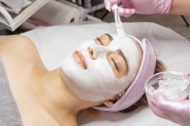 Güzellik uzmanı bir kızın yüzüne beyaz kilden bir kozmetik maske takıyor.