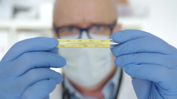 Arzt mit OP-Handschuhen und Gesichtsmaske hält ein medizinisches Thermometer in der Hand, das zur Kontrolle der menschlichen Körpertemperatur verwendet wird — Stockvideo