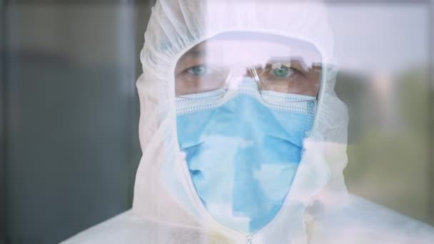 Lékař v ochranném obleku, masce obličeje, brýlích a rukavicích pro izolaci COVID-19 v zadní části okna v nemocničním pokoji — Stock video