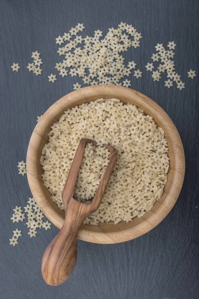 Fideos en forma de estrella en un tazón de madera y cucharada de madera de oliva sobre fondo negro de pizarra o piedra — Foto de Stock