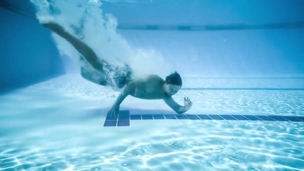 O jovem deitou-se no fundo da piscina. Sorrindo e acenando a mão debaixo de água. Movimento lento. Fotografia subaquática . — Vídeo de Stock