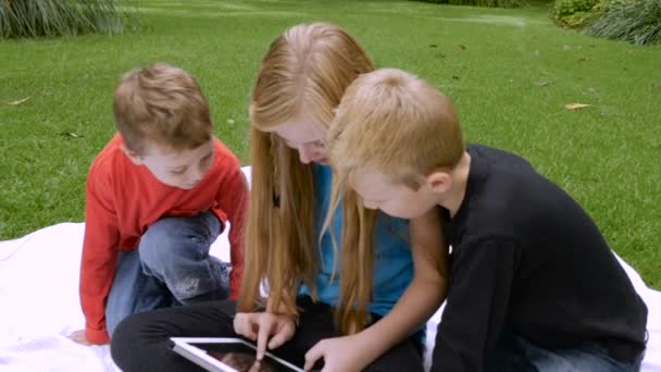Троє дітей сидять на ковдрі надворі, використовуючи планшет - повільно — стокове відео