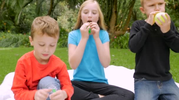 Three prepubescent children try to blow up balloons outside in yard — Stock Video