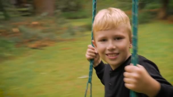 Een schattig blond haired jongen swingen op een schommel die kijken naar de camera - slowmo — Stockvideo