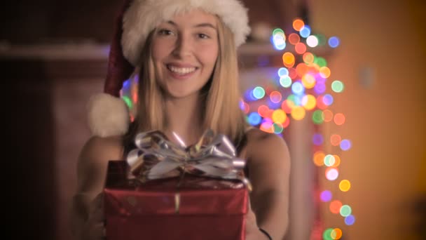 Una hermosa joven con un sombrero de Santa da un regalo envuelto en la noche — Vídeos de Stock