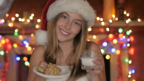 Een jonge vrouw in een kerstmuts biedt koekjes en melk rek focus op cookies — Stockvideo