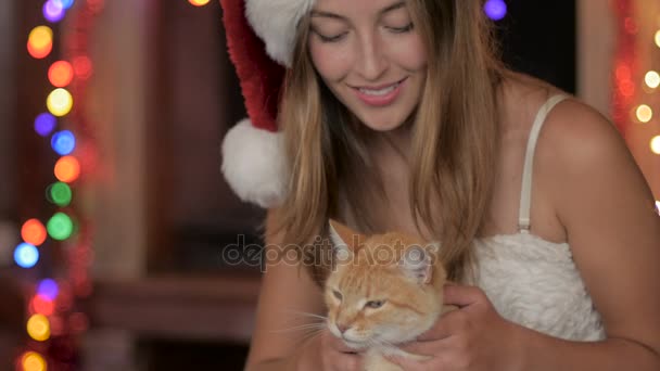 Eine lächelnde junge attraktive Frau mit Weihnachtsmütze streichelt eine orangefarbene Katze — Stockvideo