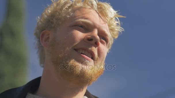 Un hombre de pelo rubio milenario barbudo sonriendo contra el cielo azul — Vídeos de Stock