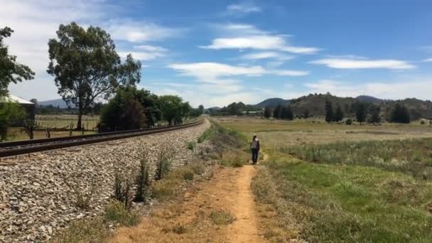 Solo mulher caminhando ao longo de uma trilha ao lado das trilhas ferroviárias em um ambiente rural — Vídeo de Stock