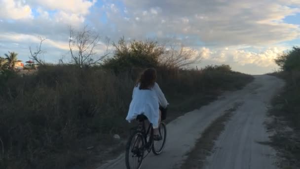 Uma mulher monta uma bicicleta na areia em direção ao pôr do sol — Vídeo de Stock
