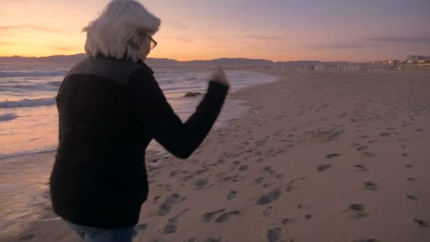 Starsza kobieta senior spaceru podekscytowany i szczęśliwy, piaszczystej plaży o zachodzie słońca — Wideo stockowe