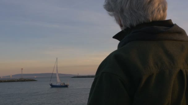 Gesunder erwachsener Mann aus den 60er Jahren, der bei Sonnenuntergang auf dem Meer oder See ein Segelboot anschaut — Stockvideo