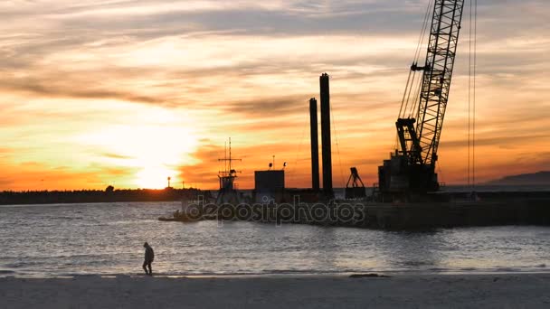 Silueta de un hombre en una sudadera con capucha caminando en la playa al atardecer con barco de draga — Vídeos de Stock