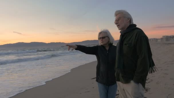 Glad mogen 60s par pekar och leende längs stranden i solnedgången — Stockvideo